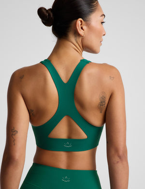 A woman shown from the back wearing a dark green athletic sports bra and matching high-waisted bottoms against a light gray studio background.