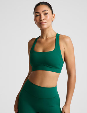 A woman modeling a dark green athletic sports bra and matching high-waisted leggings against a light gray studio background.