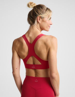 A woman modeling a matching red activewear set consisting of a sports bra and high-waisted leggings, photographed against a solid light gray studio background.