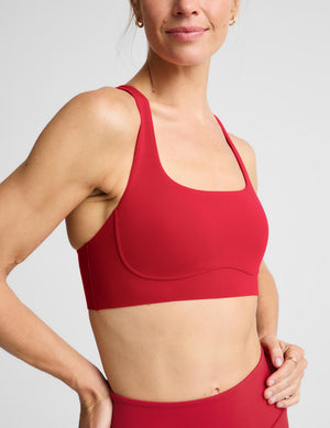 A woman modeling a matching red activewear set consisting of a sports bra and high-waisted leggings, photographed against a solid light gray studio background.