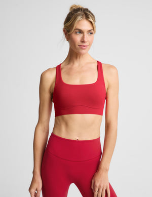 A woman modeling a matching red activewear set consisting of a sports bra and high-waisted leggings, photographed against a solid light gray studio background.