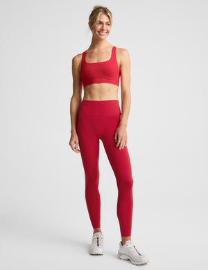 A woman modeling a matching red activewear set consisting of a sports bra and high-waisted leggings, photographed against a solid light gray studio background.