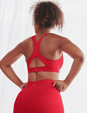 A woman shown from behind wearing a bright red sports bra and matching high-waisted leggings against a soft white curtain background.