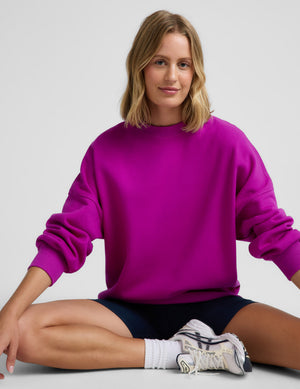 Woman wearing a purple oversized crewneck sweatshirt and navy blue biker shorts against a plain background. 
