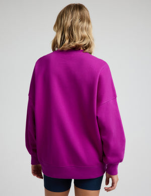 Woman wearing a purple oversized crewneck sweatshirt and navy blue biker shorts against a plain background. 