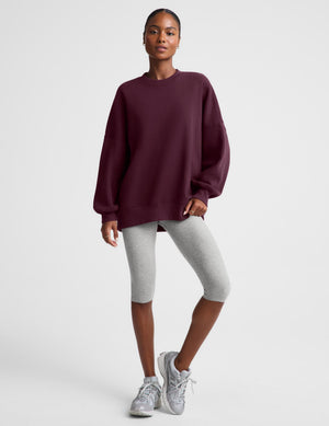 A woman modeling an oversized crewneck sweatshirt in deep burgundy, paired with light gray leggings, against a solid light gray background.