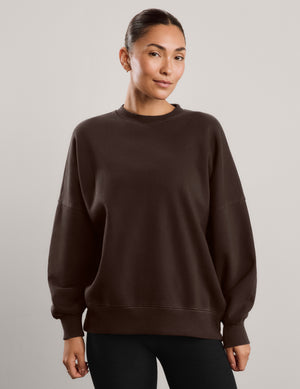 Woman wearing a brown oversized crewneck sweatshirt and black leggings against a plain background. 