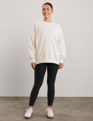 A woman wearing an oversized cream-colored crewneck sweatshirt and black leggings, against a neutral light gray studio background.