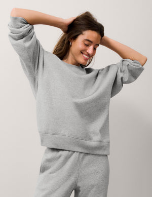 A woman wearing a light gray heathered crewneck sweatshirt and matching sweatpants, posing with her hands behind her head against a plain light gray background.