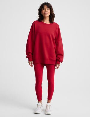 A woman wearing an oversized red crewneck sweatshirt and matching red leggings, photographed against a clean light gray studio background.