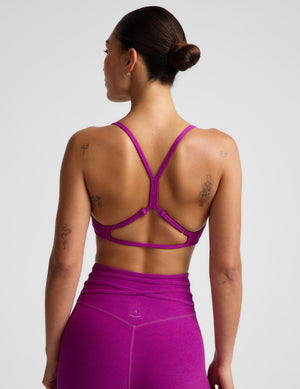 Rear view of a woman wearing a bright magenta-purple sports bra with a crisscross open-back strap design and matching high-waisted leggings against a light gray studio background.