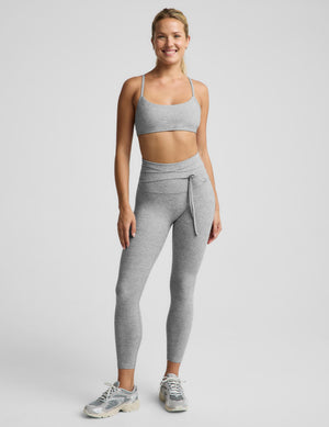 A woman modeling a light heather gray sports bra and matching high-waisted leggings with a drawstring waist, photographed against a solid light gray studio background.