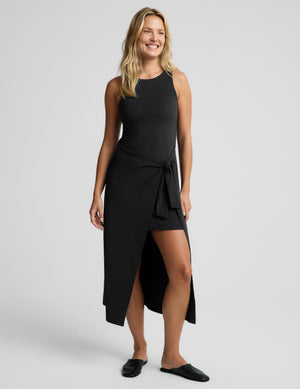 A woman modeling a sleeveless black midi dress with a high-low hem, side tie detail, and front slit, paired with black slip-on shoes against a light gray studio background.