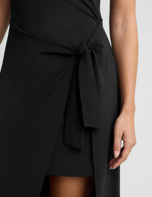 Close-up of a black wrap-style dress with a side tie knot and draped fabric, shown on a model against a light gray background.