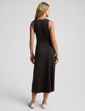 A woman viewed from the back wearing a sleeveless dark brown midi dress and light beige flats, standing against a light gray studio background.