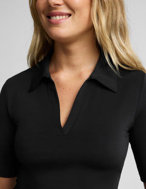 A woman modeling a black short-sleeve polo shirt with a V-neck and collar, paired with black pants, against a light gray studio background.
