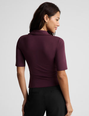 A woman wearing a short-sleeve polo shirt in deep burgundy with a split V-neckline, paired with black pants, photographed against a solid light gray studio background.
