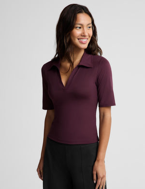 A woman wearing a short-sleeve polo shirt in deep burgundy with a split V-neckline, paired with black pants, photographed against a solid light gray studio background.