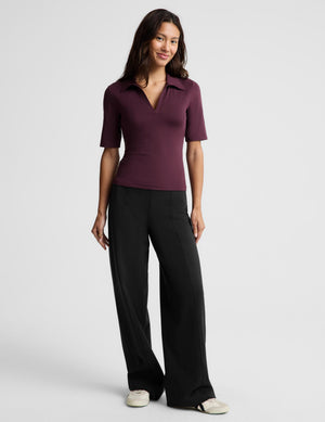 A woman wearing a short-sleeve polo shirt in deep burgundy with a split V-neckline, paired with black pants, photographed against a solid light gray studio background.