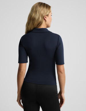 A woman shown from the back wearing a fitted short-sleeve collared top in dark navy and black leggings, photographed against a light gray studio background.