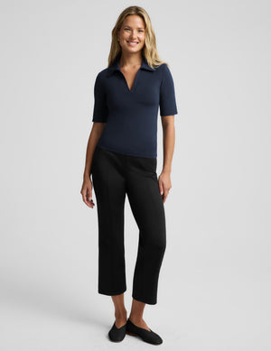 A woman modeling a short-sleeve navy polo shirt paired with black cropped pants and black flats, standing against a light gray studio background.