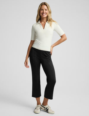 A woman modeling a fitted short-sleeve polo shirt in off-white and black cropped pants, paired with white sneakers, against a light gray studio background.