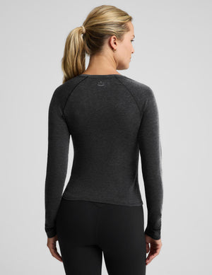 A woman with blonde hair in a ponytail is shown from the back wearing a dark heather gray long-sleeve athletic top and black leggings against a light gray studio background.