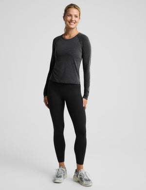 A woman modeling a dark gray long-sleeve athletic top and black leggings with gray sneakers, standing against a light gray studio background.