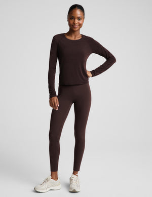 A woman modeling a dark brown long-sleeve crewneck top and matching dark brown leggings against a light gray studio background.