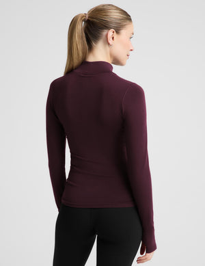 A woman modeling a fitted long-sleeve mock neck top in deep burgundy, paired with black leggings, against a solid light gray background.