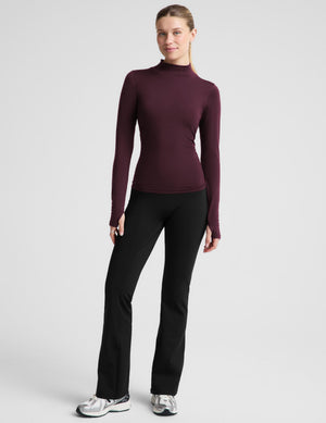 A woman modeling a fitted long-sleeve mock neck top in deep burgundy, paired with black leggings, against a solid light gray background.