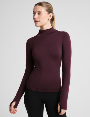 A woman modeling a fitted long-sleeve mock neck top in deep burgundy, paired with black leggings, against a solid light gray background.