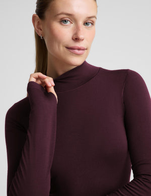 A woman modeling a fitted long-sleeve mock neck top in deep burgundy, paired with black leggings, against a solid light gray background.