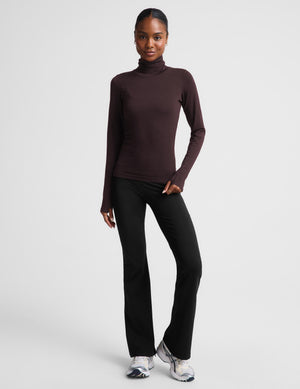 A woman modeling a dark brown long-sleeve turtle neck top and black pants against a light gray studio background.