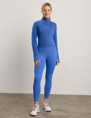A smiling blonde woman wearing a fitted royal blue long-sleeve turtle neck top and matching blue leggings, standing against a plain light gray studio background.