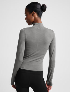 A woman modeling a fitted, long-sleeve mock neck top in a solid medium gray color, paired with black bottoms, against a light gray studio background.