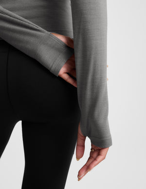 Close-up of a person wearing a gray long-sleeve top and black leggings, showing the sleeve and hand detail against a light gray background.