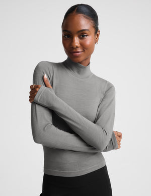A woman modeling a fitted, long-sleeve mock neck top in a solid medium gray color, paired with black bottoms, against a light gray studio background.