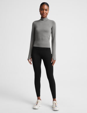 A woman modeling a fitted, long-sleeve mock neck top in a solid medium gray color, paired with black bottoms, against a light gray studio background.