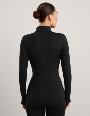 A woman modeling a black long-sleeve athletic zip-up jacket with a mock neck, paired with black leggings, against a solid light gray background.