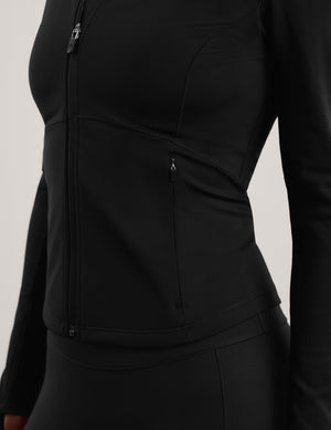A woman modeling a black long-sleeve athletic zip-up jacket with a mock neck, paired with black leggings, against a solid light gray background.