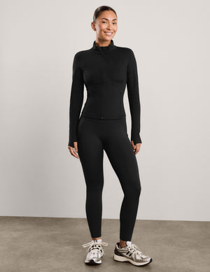 A woman modeling a black long-sleeve athletic zip-up jacket with a mock neck, paired with black leggings, against a solid light gray background.