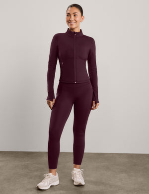 A woman modeling a long-sleeve athletic zip-up jacket and matching leggings in a deep burgundy color, set against a solid light gray studio background.