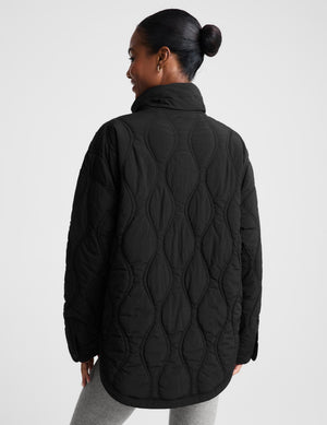 A woman wearing a black quilted zip-up jacket with a high stand-up collar, paired with light gray leggings, photographed against a plain light gray studio background.