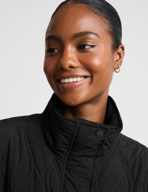 A woman wearing a black quilted zip-up jacket with a high stand-up collar, paired with light gray leggings, photographed against a plain light gray studio background.