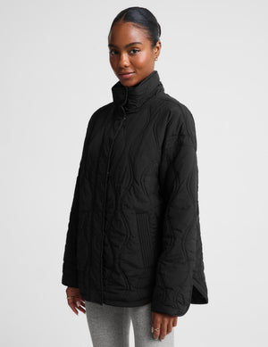 A woman wearing a black quilted zip-up jacket with a high stand-up collar, paired with light gray leggings, photographed against a plain light gray studio background.