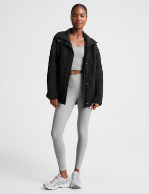 A woman wearing a black quilted zip-up jacket with a high stand-up collar, paired with light gray leggings, photographed against a plain light gray studio background.