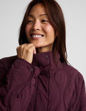A woman modeling a quilted jacket in a deep burgundy purple color, standing against a solid light gray background.