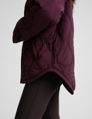 A woman modeling a quilted jacket in a deep burgundy purple color, standing against a solid light gray background.