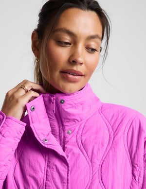 A close-up of a woman wearing a bright pink quilted zip-up jacket, adjusting the collar with her hand against a light gray background.